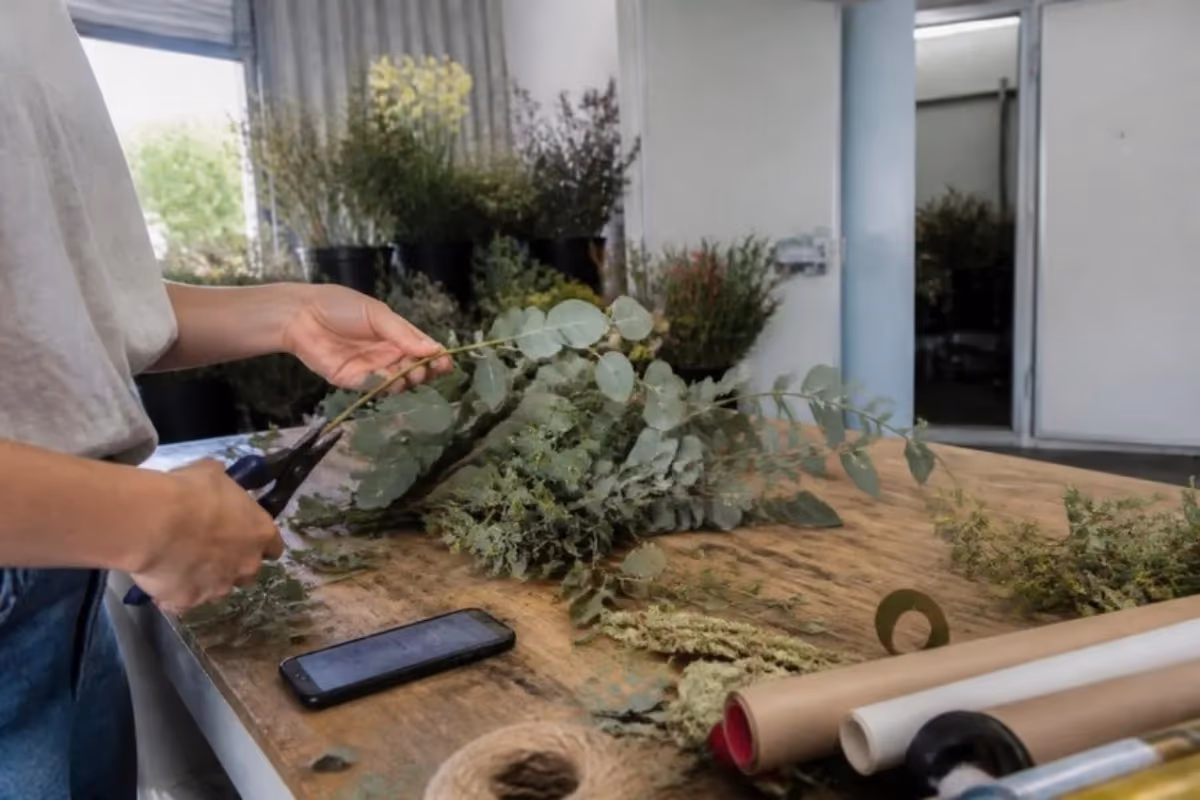 Florist trimming eucalyptus at her workbench with her phone beside her